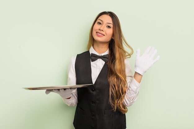 Donna graziosa asiatica sorridente felicemente agitando la mano dandoti il benvenuto e salutandoti cameriere e concetto di vassoio