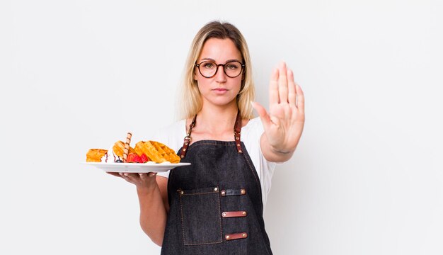 Donna graziosa bionda che sembra seria mostrando il palmo aperto che fa il concetto di cialde di gesto di arresto