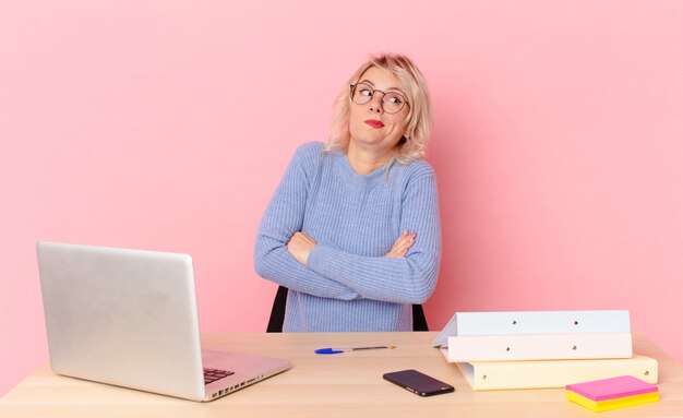 Donna graziosa bionda giovane donna graziosa che alza le spalle, sentendosi confusa e incerta. concetto di scrivania dell'area di lavoro