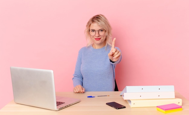 Donna graziosa bionda giovane donna graziosa che sorride e che sembra amichevole, mostrando il numero uno. concetto di scrivania dell'area di lavoro