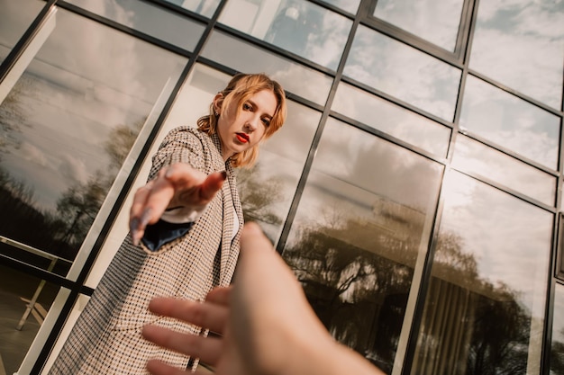 Donna graziosa che posa vicino all'edificio di vetro. Ragazza vestita abiti casual con labbra rosse che allungano la mano