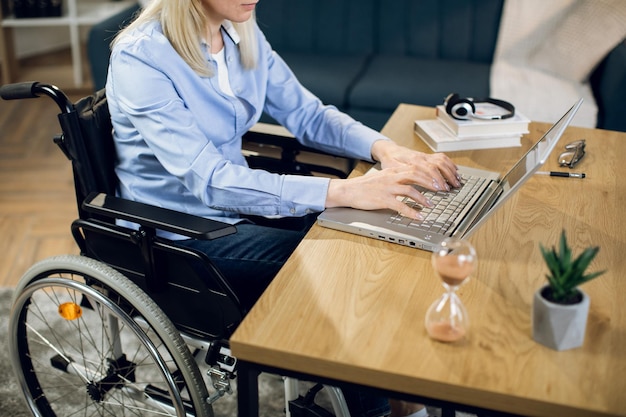Donna graziosa che si siede in sedia a rotelle e che lavora al computer portatile