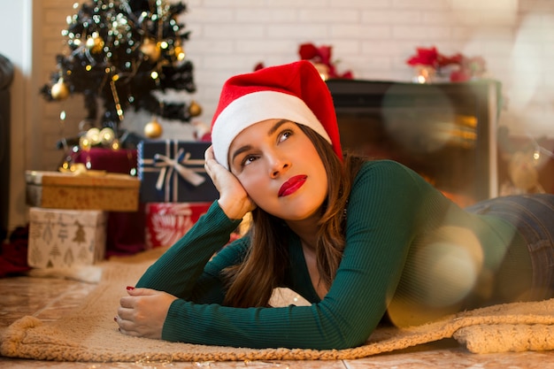 Donna graziosa che si trova sul tappeto nel salotto di casa sua con decorazioni natalizie e regali nell'albero.