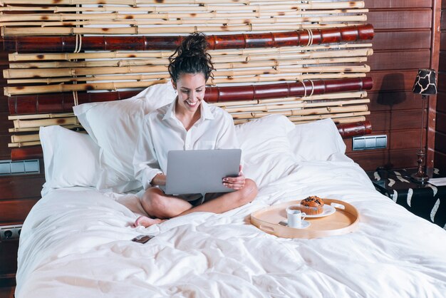 Donna graziosa con il telefono e il computer portatile a letto