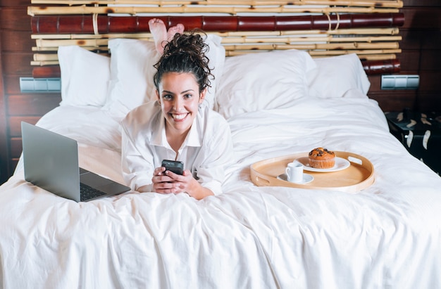 Donna graziosa con il telefono e il computer portatile a letto