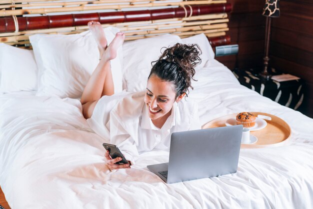 Donna graziosa con il telefono e il computer portatile a letto