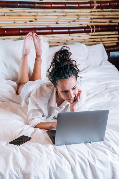 Donna graziosa con il telefono e il computer portatile a letto