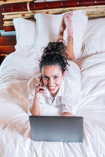 Donna graziosa con il telefono e il computer portatile a letto