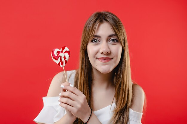 Donna graziosa con la camicetta bianca da portare della caramella del lollypop a forma di cuore isolata sulla parete rossa