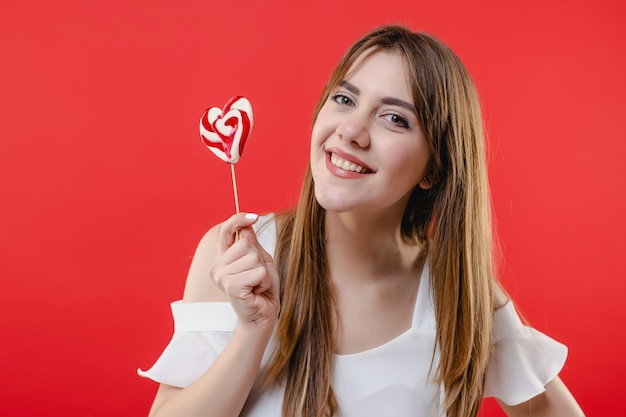 Donna graziosa con la camicetta bianca da portare della caramella del lollypop a forma di cuore isolata sulla parete rossa