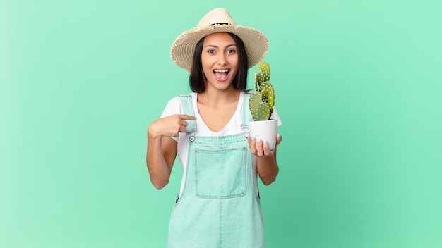 Donna graziosa dell'agricoltore che si sente felice e indica se stessa con un eccitato e tiene in mano un cactus