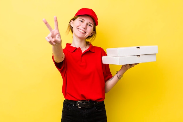 Donna graziosa della testa rossa che sorride e che sembra felice che gesturing la vittoria o il concetto della pizza di consegna della pace