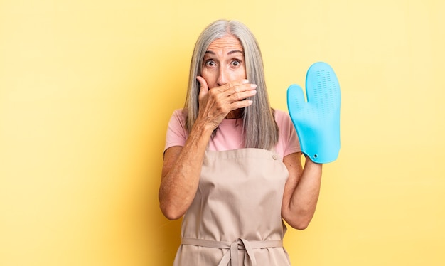 Donna graziosa di mezza età che copre la bocca con le mani con uno scioccato. concetto di guanto da forno