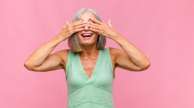 Donna graziosa di mezza età che sorride e si sente felice, coprendosi gli occhi con entrambe le mani e aspettando un'incredibile sorpresa