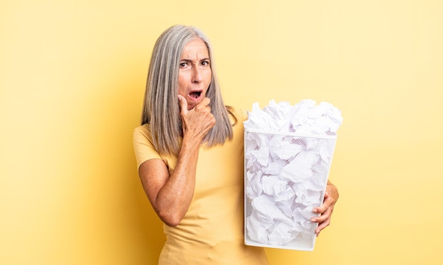 Donna graziosa di mezza età con la bocca e gli occhi spalancati e la mano sul mento. concetto di fallimento delle palle di carta