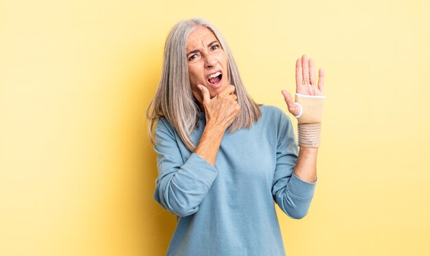 Donna graziosa di mezza età con la bocca e gli occhi spalancati e la mano sul mento. concetto di fasciatura per le mani