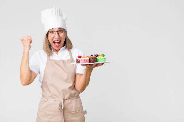 Donna graziosa di mezza età del panettiere con le torte contro la parete