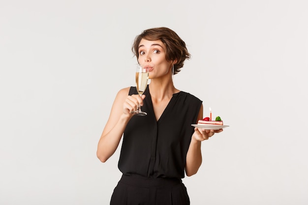 Donna graziosa divertente sorseggiando champagne e tenendo un pezzo di torta di compleanno, in piedi sopra il bianco.