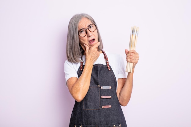 Donna graziosa senior con la bocca e gli occhi spalancati e la mano sul mento. artista con i pennelli