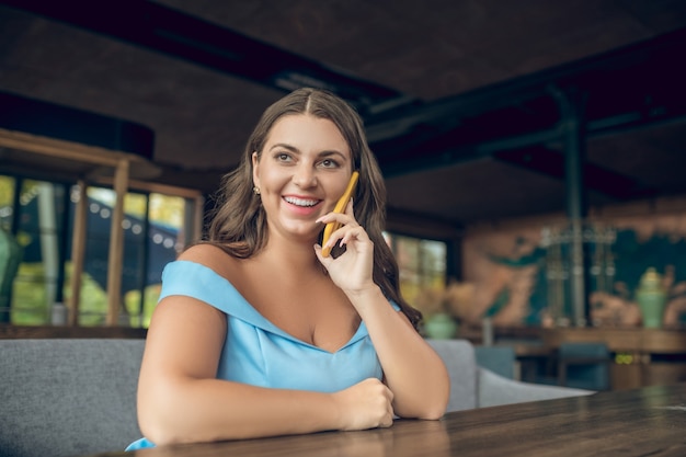 Donna graziosa sorridente in vestito blu con lo smartphone vicino all'orecchio che si siede al tavolo in caffè estivo di ottimo umore