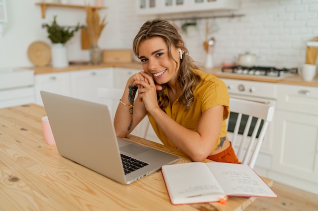 Donna graziosa studentessa che usa il computer portatile e scrive appunti sulla sua moderna cucina leggera.