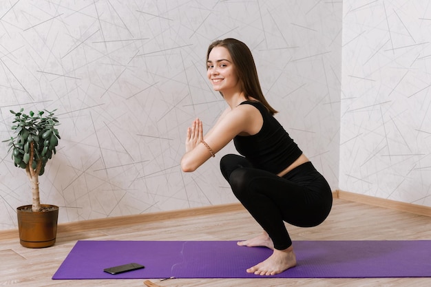 donna in abbigliamento sportivo nero che fa yoga in posa Garland mentre è seduto sul tappetino