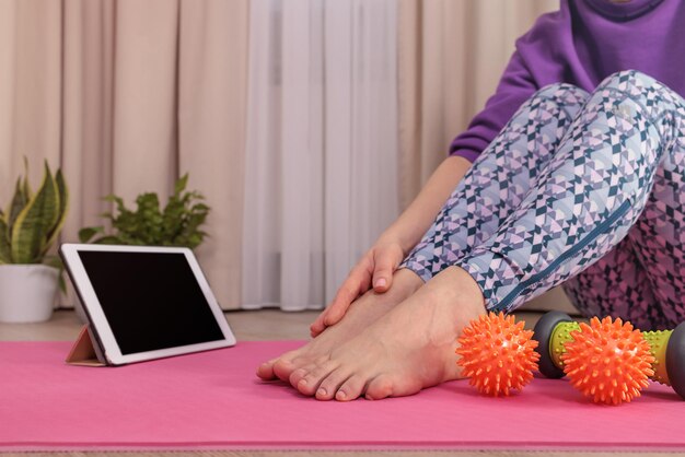 Donna in abbigliamento sportivo Veri Perry seduta su un tappetino da yoga rosa che guarda il tablet che massaggia le gambe a casa