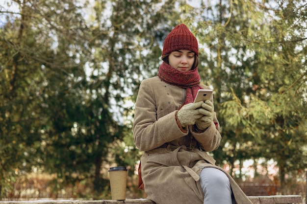 donna in abiti caldi che naviga sul cellulare mentre si siede sulla panchina nel parco invernale innevato