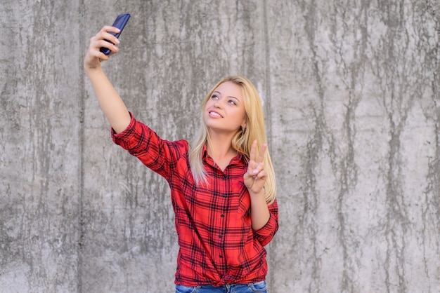 Donna in abiti casual con un sorriso raggiante che racconta selfie sul suo smartphone e mostra il v-sign