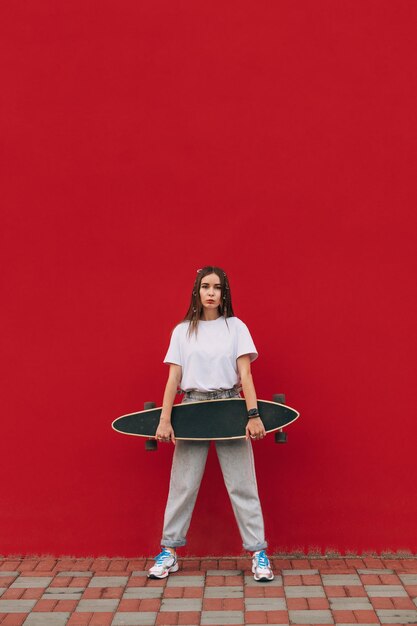 donna in abiti eleganti in posa su uno sfondo di un muro rosso con un longboard nelle sue mani