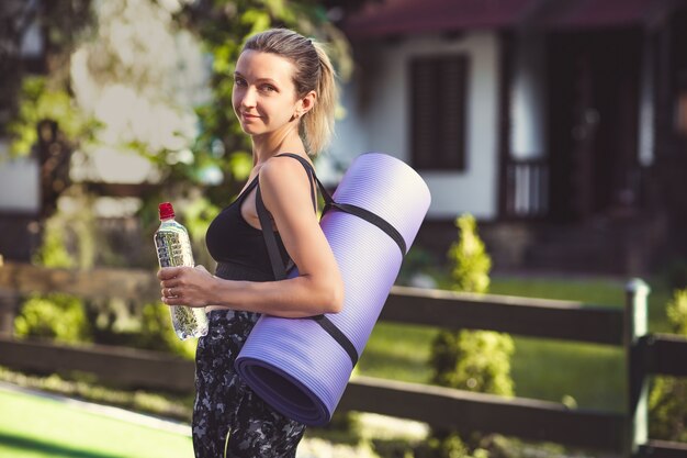 Donna in abiti sportivi in possesso di un materassino yoga e una bottiglia d'acqua dopo l'allenamento.