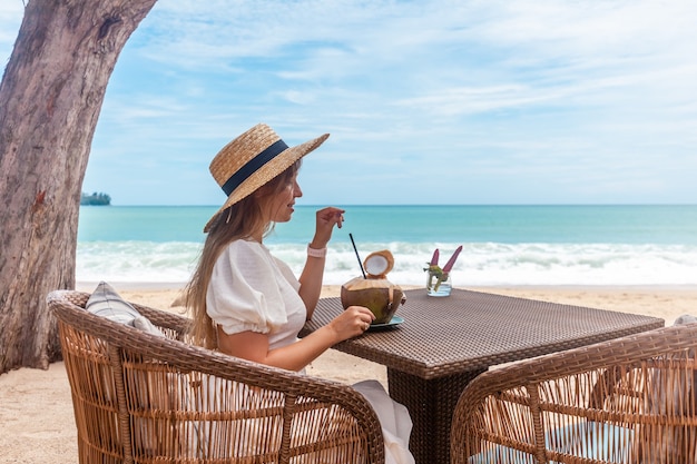 Donna in abito bianco e cappello di paglia seduto sul tavolo in un caffè all'aperto sulla spiaggia