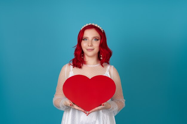 donna in abito bianco e con i capelli rossi tiene un grande cuore di carta rossa contro il petto