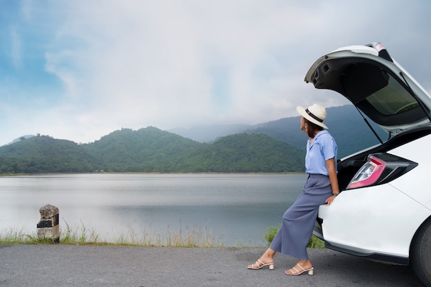 Donna in abito blu, indossa un cappello bianco, seduto nel bagagliaio di un'auto, guardando una montagna con un fiume