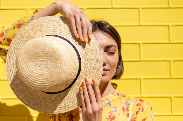 Donna in abito estivo giallo e cappello sul muro di mattoni gialli calma e positiva, gode di soleggiate giornate estive