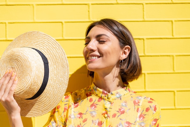 Donna in abito estivo giallo e cappello sul muro di mattoni gialli calma e positiva, gode di soleggiate giornate estive