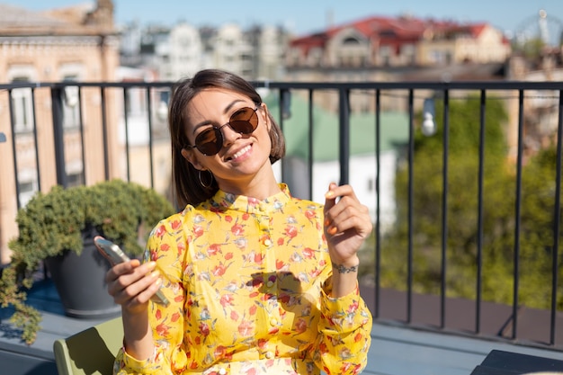 Donna in abito giallo sulla terrazza in un caffè estivo con il telefono cellulare al giorno pieno di sole, sembra felice e positiva con un enorme sorriso sul viso