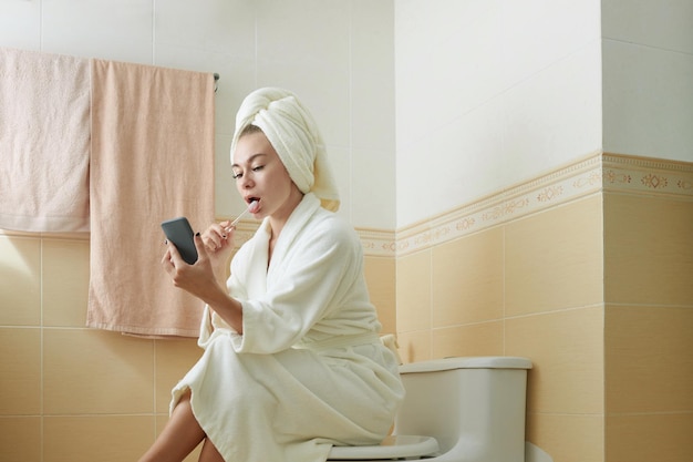 Donna in accappatoio seduta sul water che si lava i denti e legge notizie di messaggi di testo o e-mail sullo smartphone