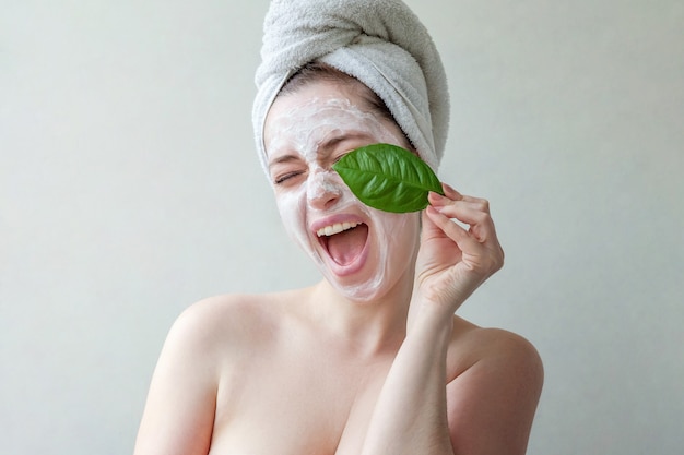 Donna in asciugamano sulla testa con maschera nutriente bianca o crema sul viso, sfondo bianco