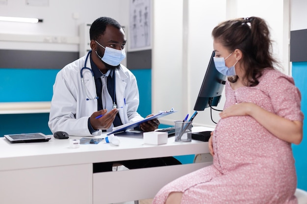 Donna in attesa di un bambino e che discute con il medico sulla gravidanza e sull'assistenza sanitaria. Medico di medicina generale che fornisce consulenza medica a pazienti in gravidanza durante la pandemia di coronavirus