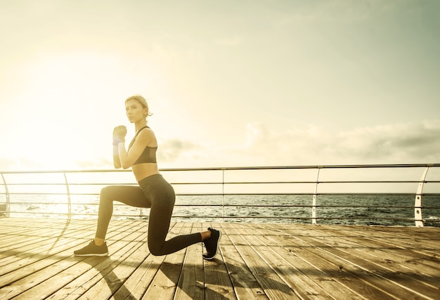 Donna in buona salute formazione sul lungomare. La giovane donna attraente in abiti sportivi fa esercizio di affondi sulla spiaggia all'alba. Riscaldamento