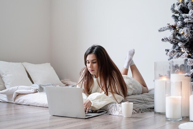 Donna in caldo maglione invernale bianco sdraiato a letto a casa utilizzando il computer portatile