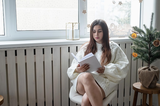 Donna in caldo maglione invernale bianco seduto accanto alla finestra a casa alla vigilia di Natale leggendo il libro