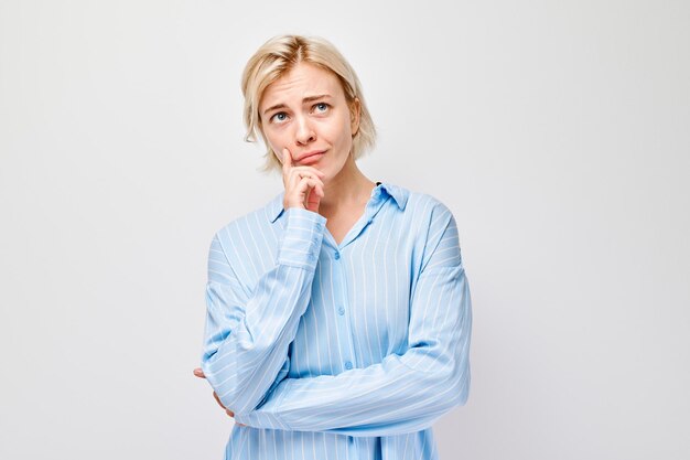 Donna in camicia blu che sembra perplessa con la mano sul mento sullo sfondo bianco