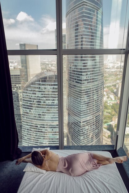 Donna in camicia da notte sdraiata sulla finestra del letto in un grattacielo vicino al letto