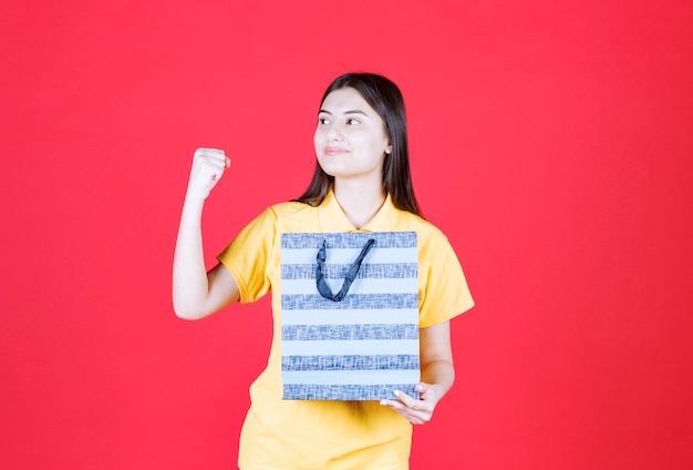 donna in camicia gialla che tiene un sacchetto della spesa blu e che mostra il segno positivo della mano.