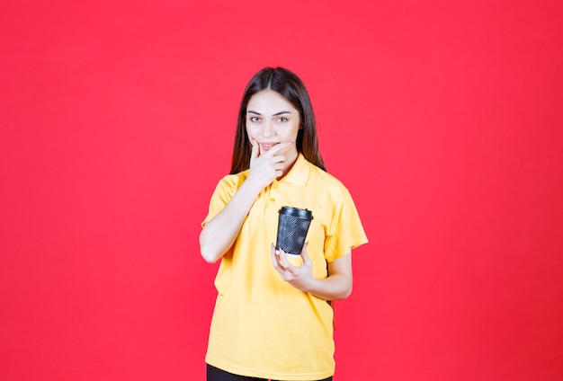 donna in camicia gialla che tiene una tazza di caffè usa e getta nera, pensando e avendo una buona idea.