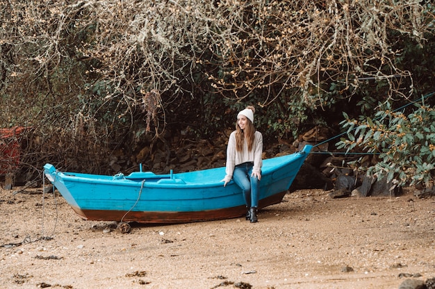 Donna in cappello e maglione che si siede sulla vecchia barca di legno ormeggiata sulla riva sabbiosa in autunno e distoglie lo sguardo