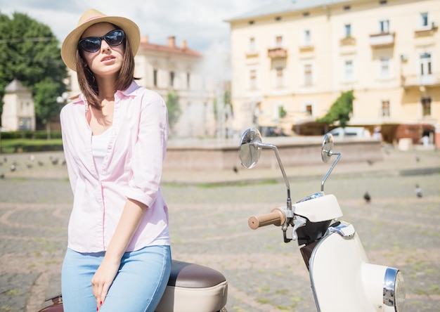 Donna in cappello elegante che sta motorino vicino all'aperto.