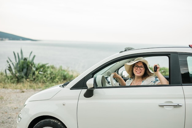 Donna in cappello estivo che tiene le chiavi della macchina alla guida della sua nuova automobile cabriolet e concetto di acquisto